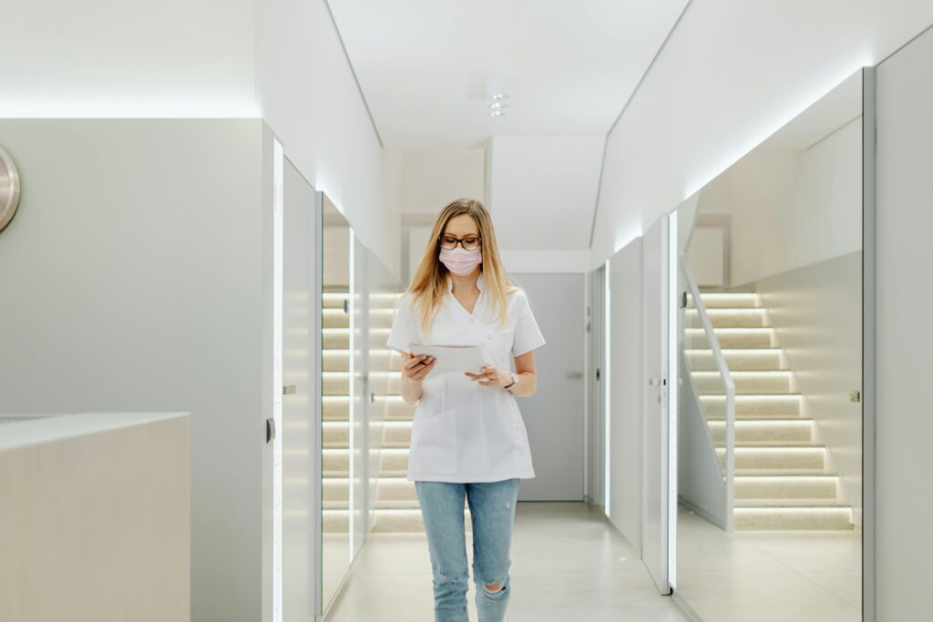 Masked woman in casual attire walking down a bright modern hallway holding a tablet.