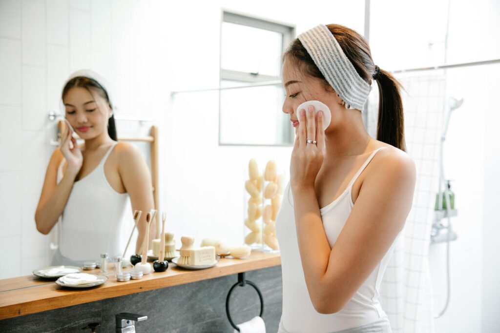 Asian woman applying skincare with cotton pad in bright bathroom setting.