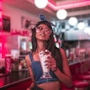 Young woman in fashionable attire enjoying a milkshake at a retro diner with vibrant lighting.