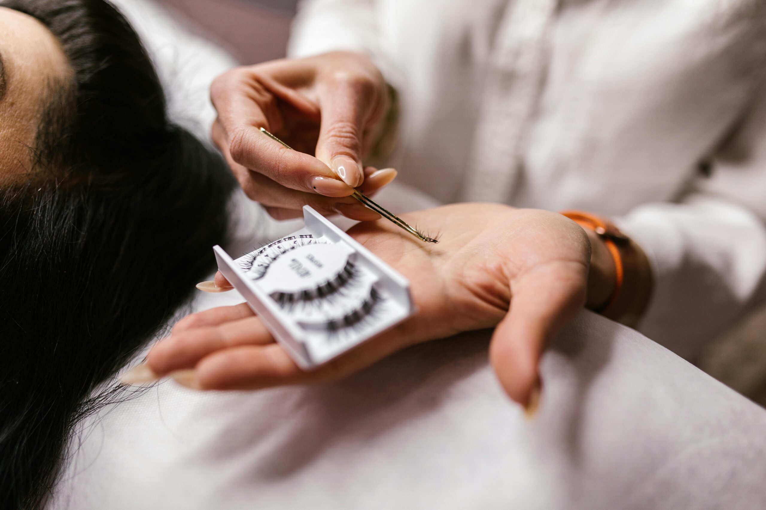 home - beauty directory - Close-up of an eyelash extension application using tweezers, featuring a beautician's hands.