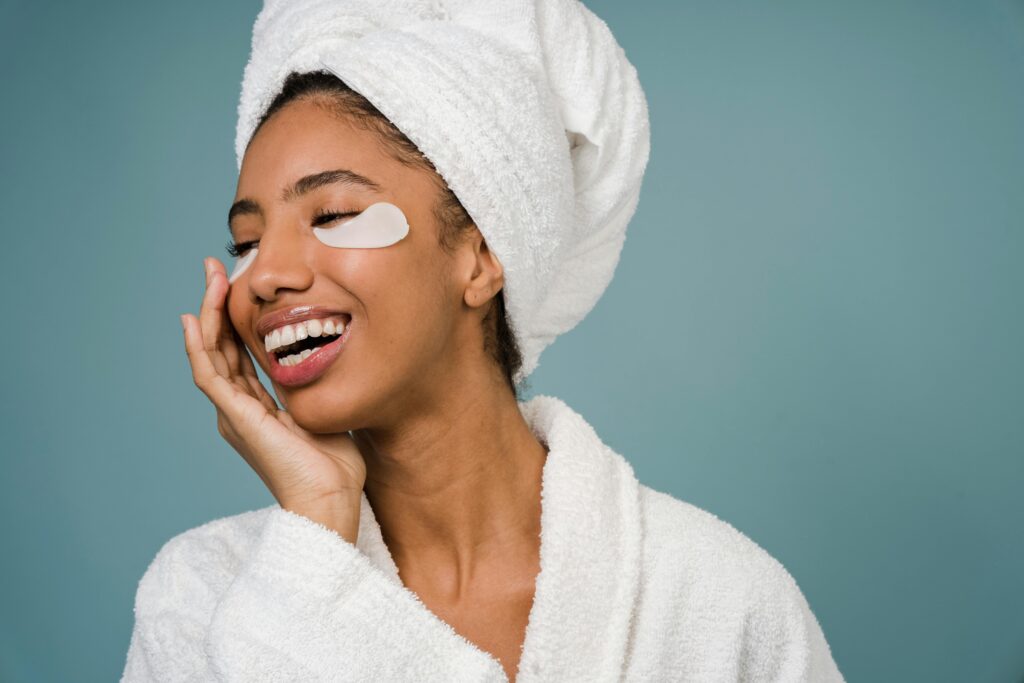 Smiling woman in a bathrobe with eye patches, promoting skincare and wellness.